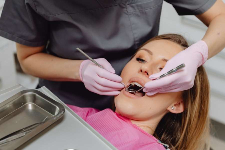 Un dentiste en blouse grise et gants roses examine les dents d'une patiente avec un miroir.