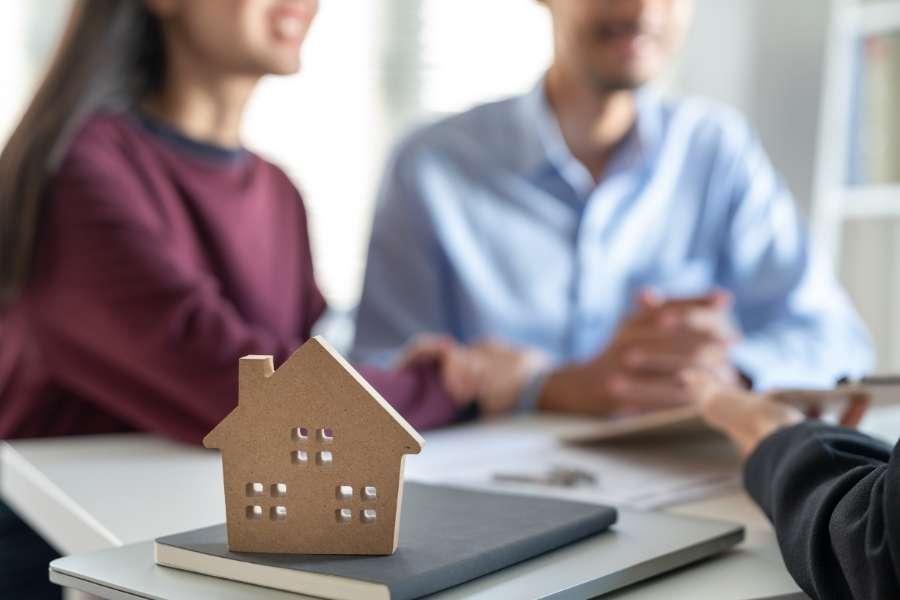 Un couple est assis en face d'un conseiller avec une maquette de maison en bois au premier plan, évaluant la rentabilité réelle d'un bien immobilier.