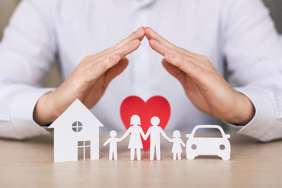 Un homme en chemise blanche protège avec ses mains une famille en papier, une maison, une voiture et un cœur rouge sur une table en bois.